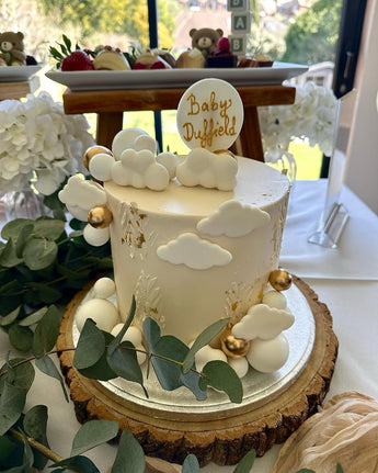 White baby shower cake with clouds, gold balls and a Baby topper on a wooden slice