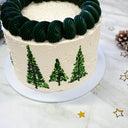 White Christmas Tree Cake with dark green piped trees and swirls, with a pine cone and gold stars