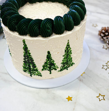 White Christmas Tree Cake with dark green piped trees and swirls, with a pine cone and gold stars
