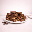 Wholesale Tiffin bars on a plate, showcasing smooth chocolate tops with nut-studded fillings. Extra chocolate pieces on the side enhance the indulgent presentation.