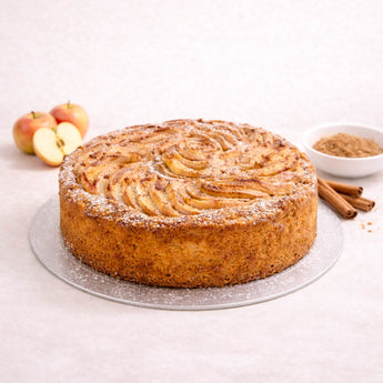 Dorset Apple Cake topped with sliced apples and icing sugar, next to apples and cinnamon sticks