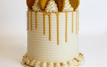 A tall round cake with white frosting, caramel drip, topped with cookie-decorated swirls, and bordered by frosting rosettes, showcasing Harry Batten's bespoke craftsmanship.