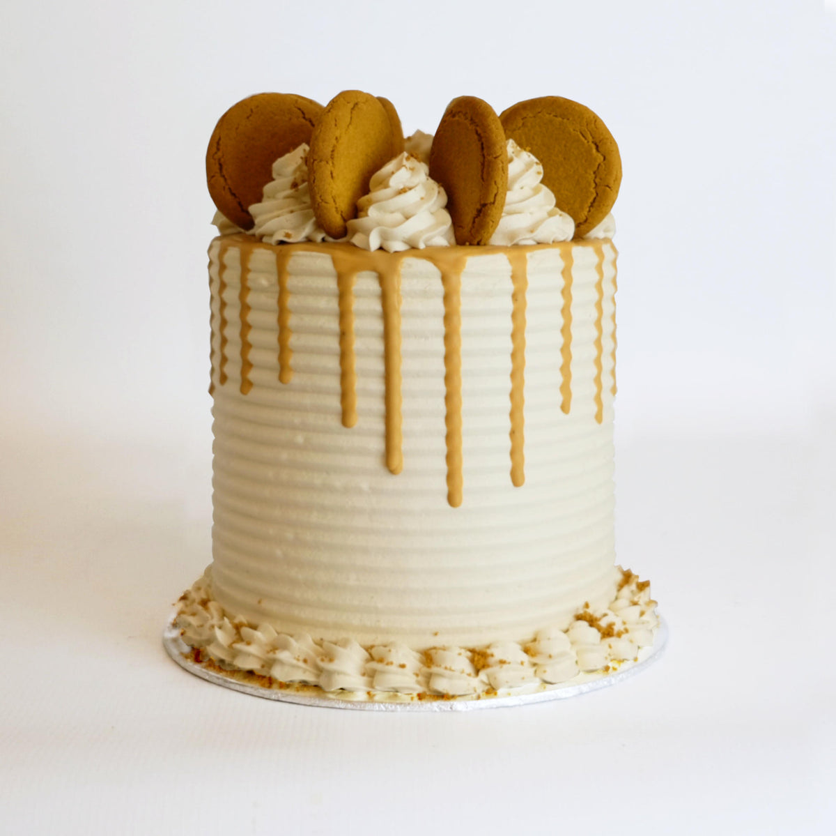 A tall round cake with white frosting, caramel drip, topped with cookie-decorated swirls, and bordered by frosting rosettes, showcasing Harry Batten's bespoke craftsmanship.