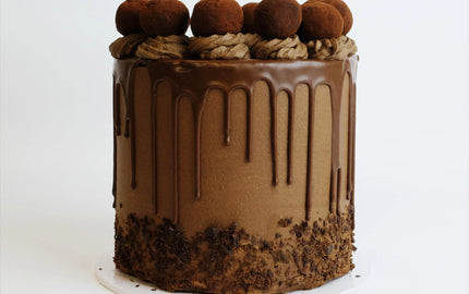 A tall, round chocolate cake with chocolate buttercream, ganache drip, and cocoa-dusted truffles, topped with chocolate frosting swirls and shavings at the base.