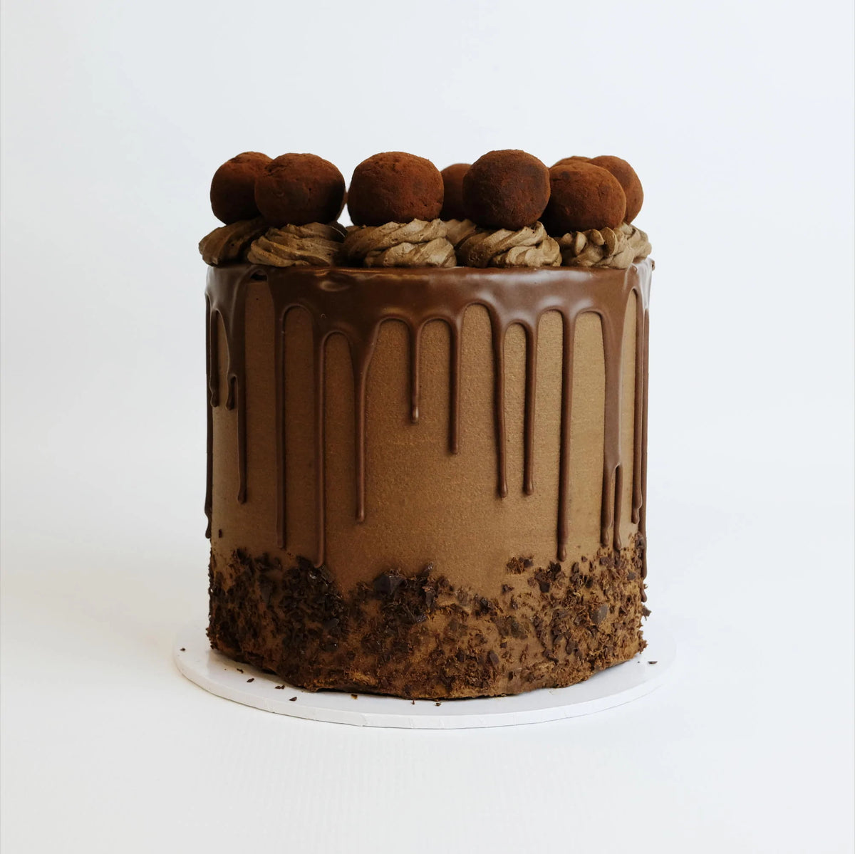 A tall, round chocolate cake with chocolate buttercream, ganache drip, and cocoa-dusted truffles, topped with chocolate frosting swirls and shavings at the base.
