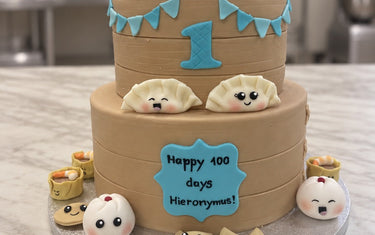 Two-tier dim sum cake styled as bamboo steamers with smiling dumplings, blue bunting and a Happy 100 days plaque