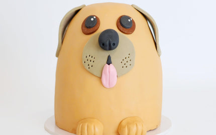 Cartoon-style dog cake with orange fondant, featuring floppy ears, big eyes, snout with tongue, and front paws; a creative novelty by Harry Batten.