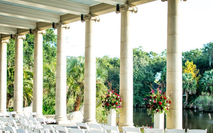 Outdoor pavilion set for a wedding with white chairs, floral arrangements, and classical columns overlooking a pond and lush greenery.