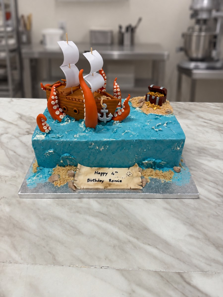 Blue buttercream sea cake with a sugar pirate ship, orange kraken tentacles, a treasure chest island and birthday scroll