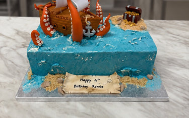 Blue buttercream sea cake with a sugar pirate ship, orange kraken tentacles, a treasure chest island and birthday scroll