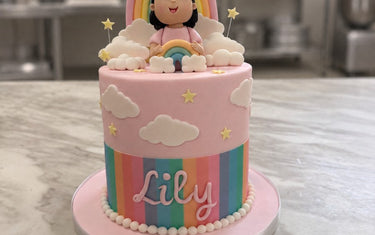Pink rainbow birthday cake with clouds, stars, pastel stripes, a sugar girl topper holding a rainbow and Lily in pink script