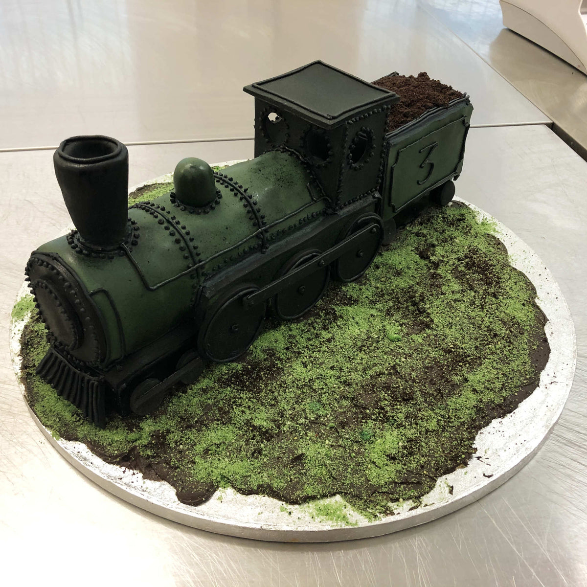 Novelty cake styled as a vintage steam locomotive in muted green, with chocolate “coal” in tender, on grass and dirt-like cake board.