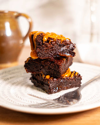 Stack of chocolate brownies on a white plate, drizzled with caramel sauce, accompanied by a silver fork; ceramic mug blurred in the background.
