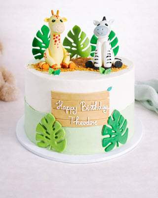 Jungle-themed birthday cake with giraffe and zebra figurines, tropical leaves, and a Happy Birthday Theodore plaque on pale green and white frosting.