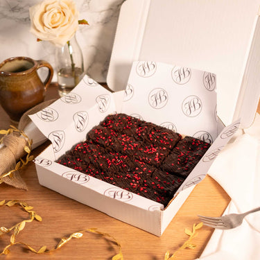 Open box of Raspberry and Amaretti Brownies with chocolate drizzle, next to a mug and a rose