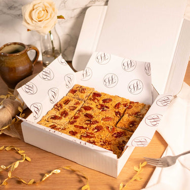 Open box of Maple Pecan Blondies topped with pecans, with a rose and a mug