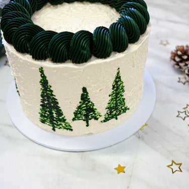 White Christmas Tree Cake with dark green piped trees and swirls, with a pine cone and gold stars