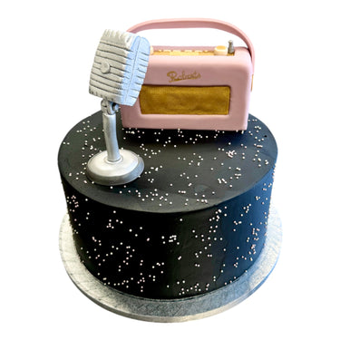 Custom Cake featuring matte black buttercream, tiny white sugar pearls, a vintage-style microphone, and retro radio fondant decorations atop a silver cake board.