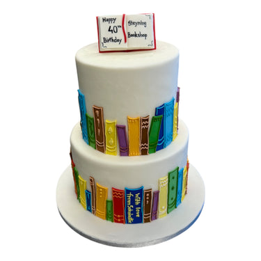 Custom cake with two tiers, white fondant, rainbow book spines, and an open book topper inscribed “Happy 40th Birthday Steyning Bookshop.”