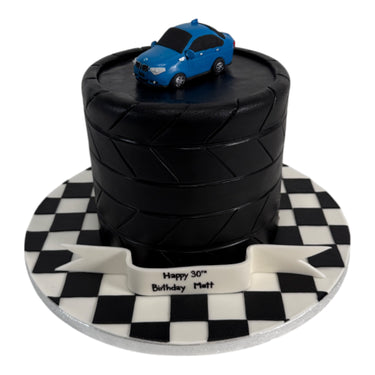 Custom Cake: Tire-themed birthday cake with black fondant, blue toy car, and checkered board. Banner reads “Happy 30th Birthday Matt.” Contact us before purchasing.