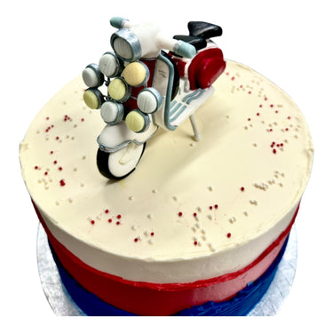 Custom Cake with frosted blue, red, and ivory bands, topped with red and pearl sprinkles and a scooter cake-topper. Contains gluten, milk, egg, nuts, and soya.