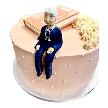 Custom Cake featuring a pale-pink frosting with white pearls and buttercream swirls, adorned with an edible elderly woman figurine and a fondant book model.
