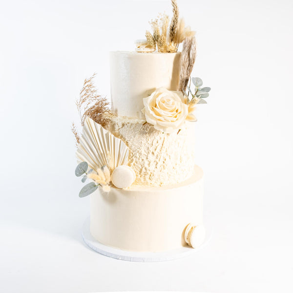 Boho Wedding Cake with ivory buttercream, adorned with a sugar-paste rose, dried grasses, bleached palm leaf, and white macarons, showcasing a minimalist, elegant design.