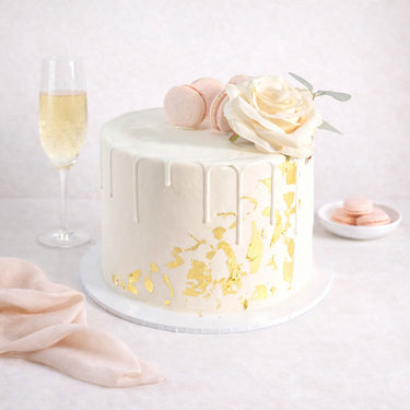 Gold Leaf Cake with white buttercream, gold leaf, macarons and a rose, next to a glass of fizz