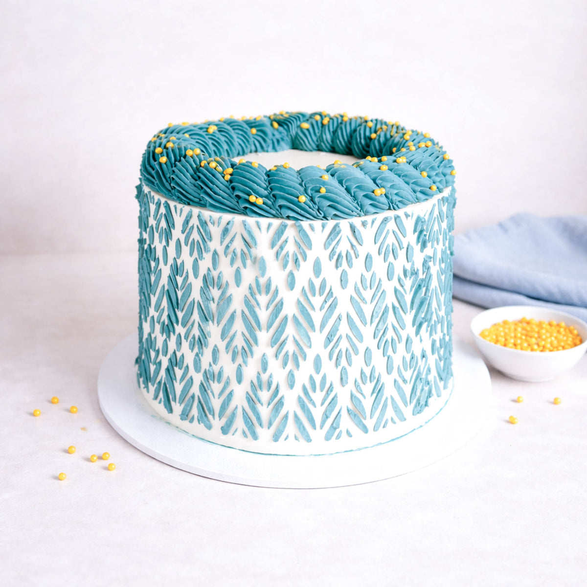 Stencil Cake with a teal leaf pattern and yellow sugar pearls, on a white cake board