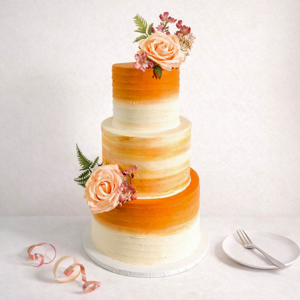 Three-tiered Ombré Wedding Cake with smooth buttercream in white to warm orange shades, adorned with blush-peach roses and greenery, on a white tabletop.