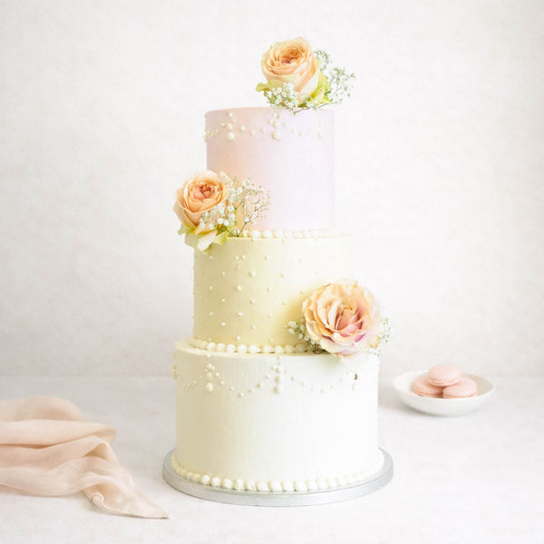 Three-tier Formal Wedding Cake with piped pearls, fresh roses, and baby's-breath, accompanied by pink macarons and a pale pink cloth.