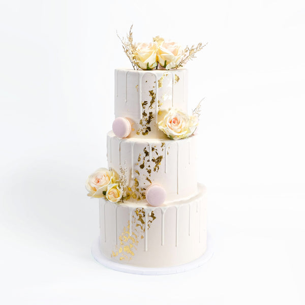 Gold Leaf Wedding Cake with white drip icing, adorned with pale-cream roses, pink macarons, dried floral sprigs, and gold leaf, offering a refined elegance.