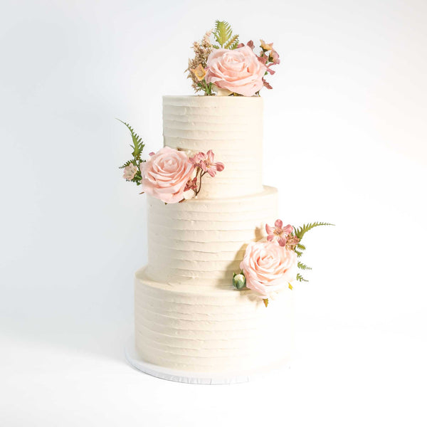 Rustic Wedding Cake with white frosting, adorned with pink sugar roses and greenery, showcasing a simple, elegant design. Perfect for personalized weddings, handmade by Harry Batten.