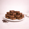 Wholesale Tiffin bars on a plate, showcasing smooth chocolate tops with nut-studded fillings. Extra chocolate pieces on the side enhance the indulgent presentation.