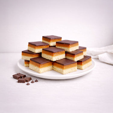 Wholesale Millionaire Slice: Three-layer bars with shortbread base, caramel middle, and chocolate top on a plate, featuring extra chocolate pieces and chips.